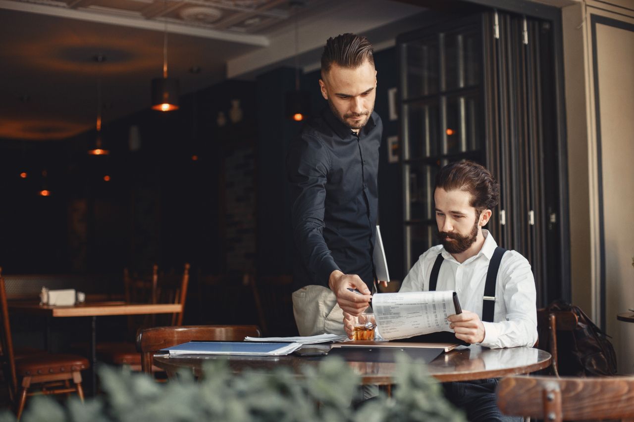 Two men discuss business over documents in a cozy, stylish restaurant.